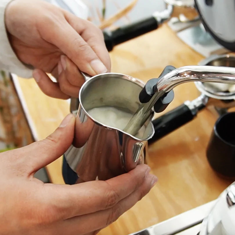 Person holding a metal milk frothing pitcher near a coffee machine on a wooden surface.