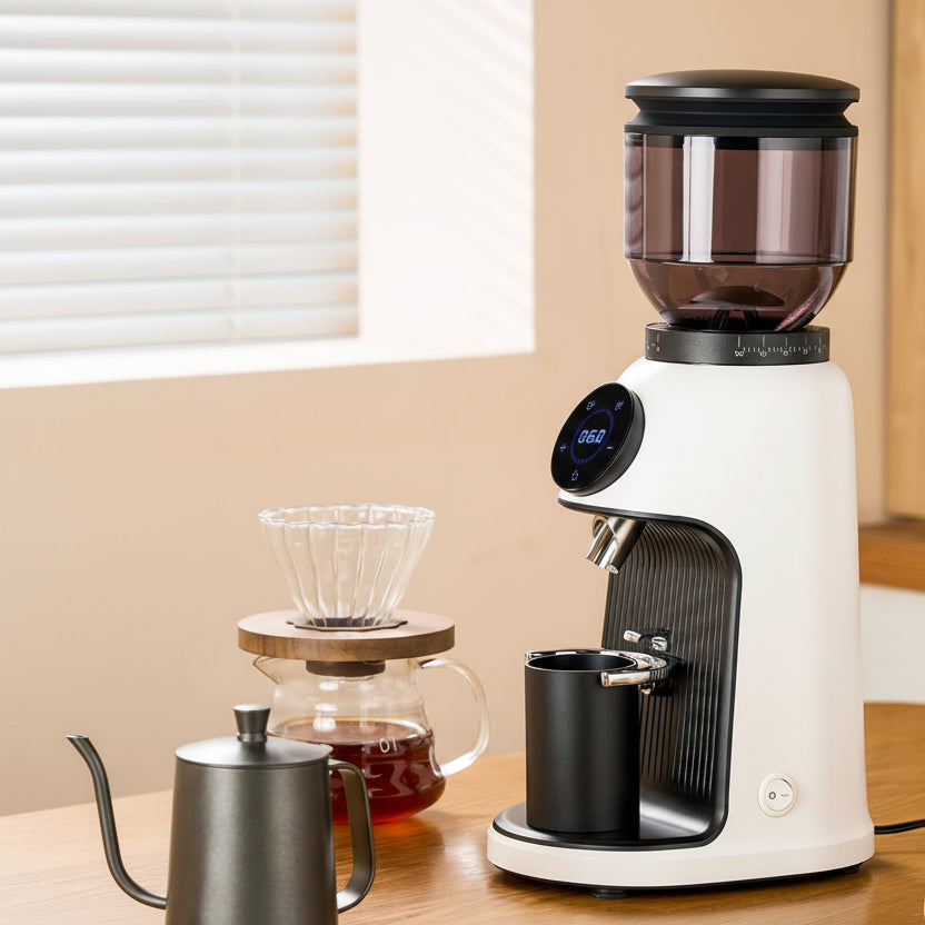 Coffee grinder on a wooden table with a server of coffee and other kitchen items.